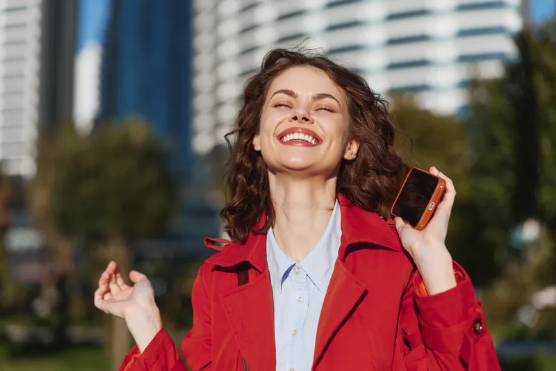 A smiling woman in a red jacket stands outdoors in the city, holding a phone near her ear, enjoying a sunny day and urban breeze.