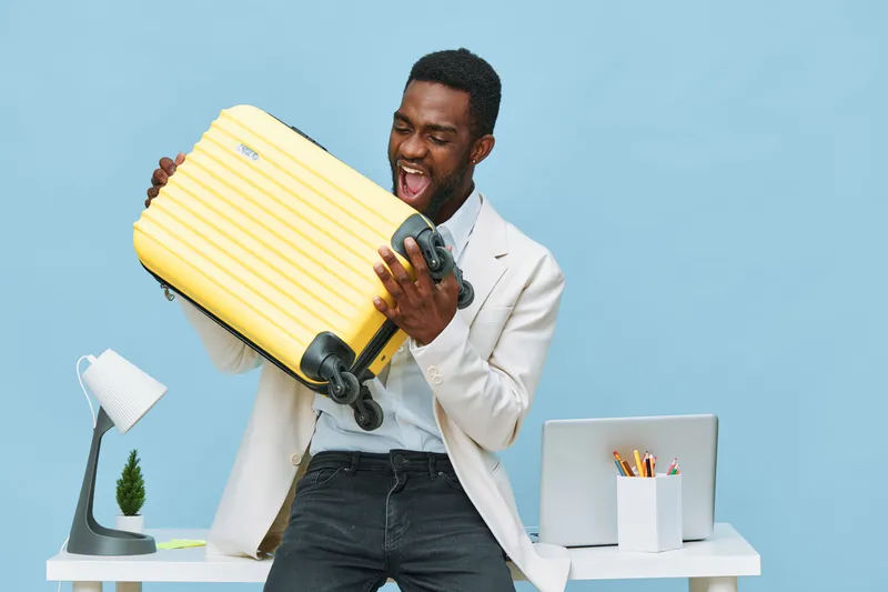 Joyful man in white blazer and dark jeans holding yellow suitcase expressing excitement in bright studio setup with modern office desk and tablet concept: travel adventure adventure happiness