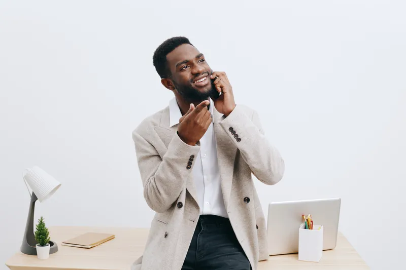 Confident smiling black man talking on mobile phone in modern office with laptop, desk accessories, and bright minimalistic interior style. Business communication, technology, professional lifestyle c