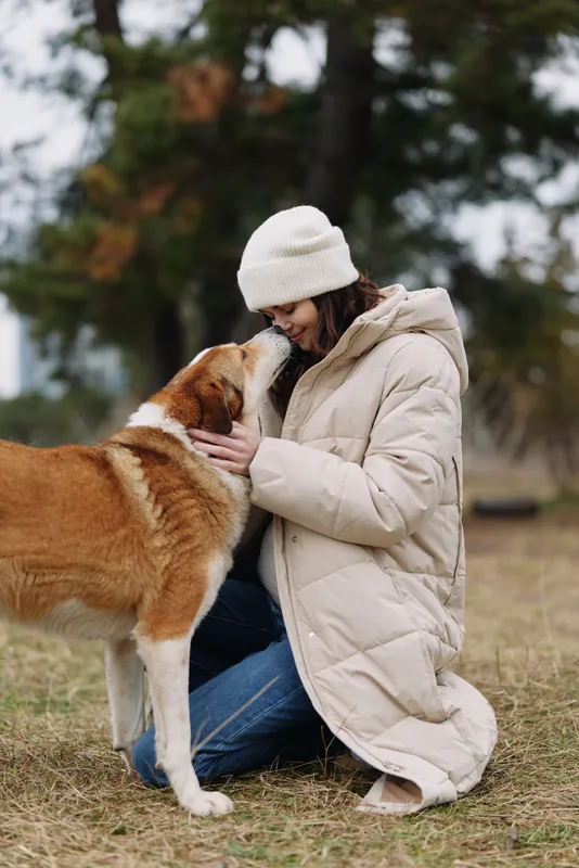 A person wearing a warm beige coat and knit hat kneels on a field, sharing a tender moment with a fluffy dog, expressing companionship and cold weather comfort.