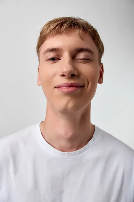 Close up of a young man with a playful wink and a bright smile, wearing a white t shirt, casual expression in a clean studio setting, showcasing friendly mood and approachable charm