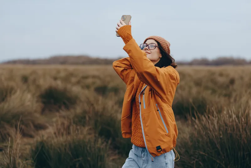 Woman outdoors lifestyle field hat sunlight smile autumn, cheerful woman in a wide grass field lifting a straw hat, tan jacket and jeans, relaxed pose, gentle wind, natural colors