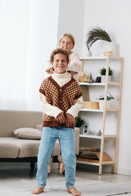 Two smiling children pose in a bright living room, the older girl behind the boy, hands on his shoulders, wearing a knit vest and jeans, framed by soft light.
