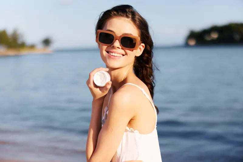 Happy woman wearing sunglasses at the seaside holding a seashell, sunlit portrait with calm water and distant shoreline, summer mood and carefree vibe.