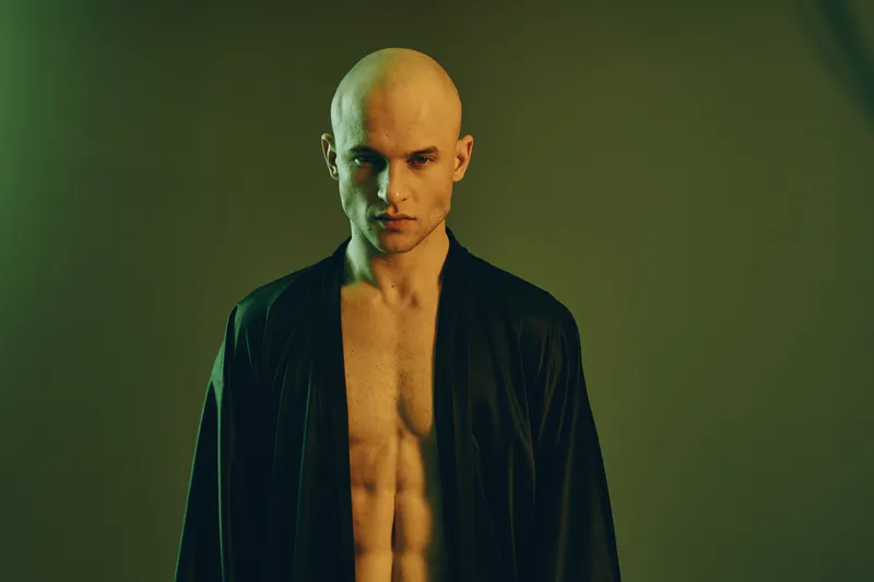 Intense man posing confidently in a stylish black robe, showcasing a muscular physique with dramatic lighting in a moody green atmosphere.