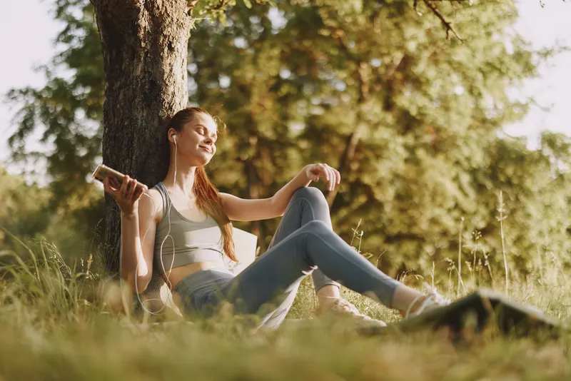 Young woman enjoying music outdoors in a serene park, wearing casual sportswear, exuding happiness in a sunlit natural setting