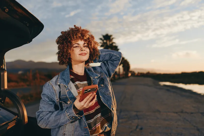 Woman lifestyle outdoors at sunset, curly hair, denim jacket, holding smartphone near open car trunk, candid travel moment, warm golden sky, road and ocean horizon, soft backlight