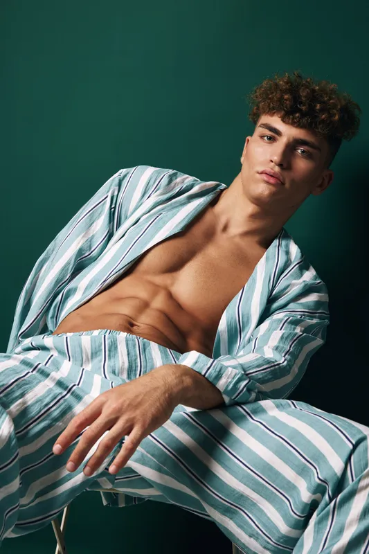 Young man with curly hair wearing striped open shirt and pants posing against green background with serious expression and relaxed posture in studio.