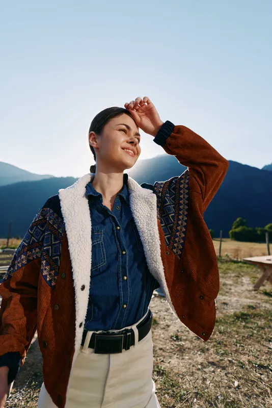 Woman with a warm smile wearing a patterned jacket and denim shirt, portrait in sunlight against mountains outdoors, standing in a field enjoying nature and relaxed travel style.