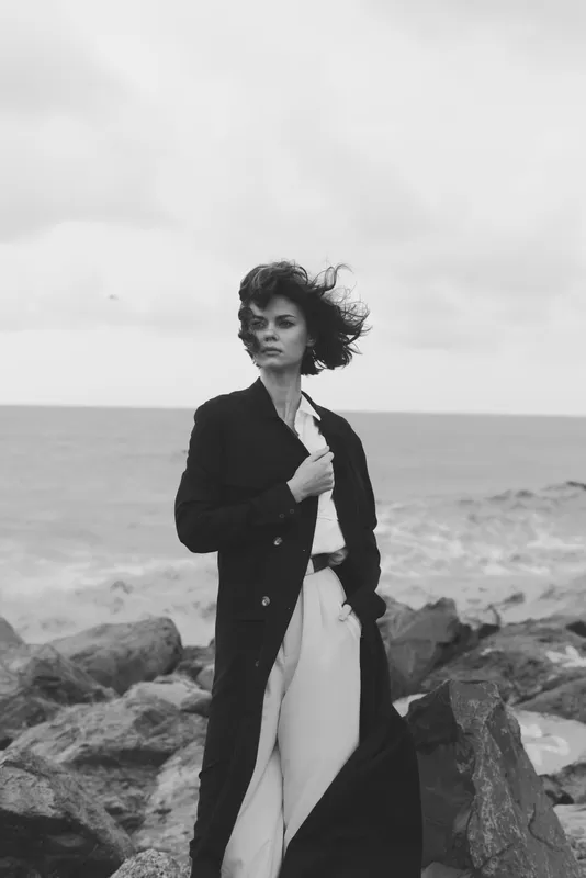 Woman on a windswept beach wearing a long coat and flowing trousers, standing among rugged rocks by the sea, monochrome scene, moody atmosphere, fashion photography, natural light, candid pose,
