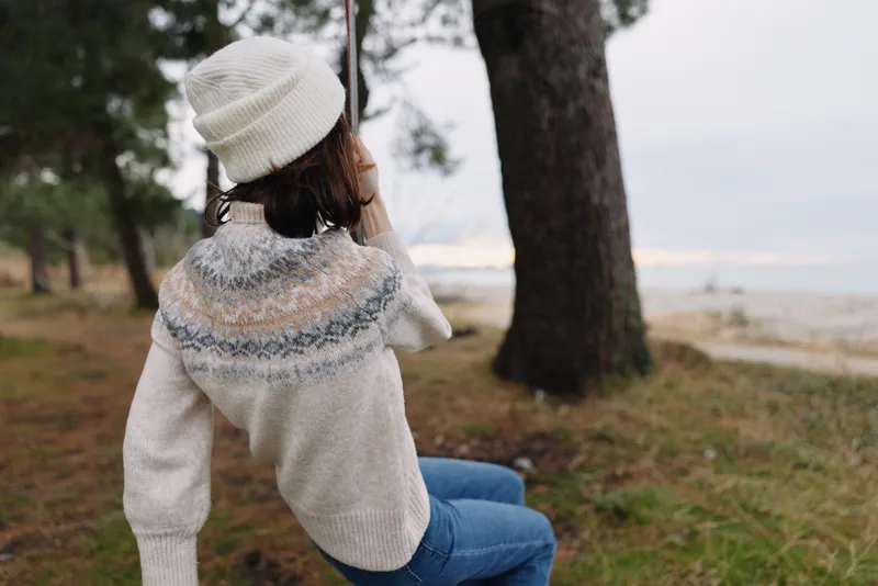 Outdoor woman in a cozy sweater and beanie sits by a lake, trees frame the calm water and distant horizon in a serene natural scene