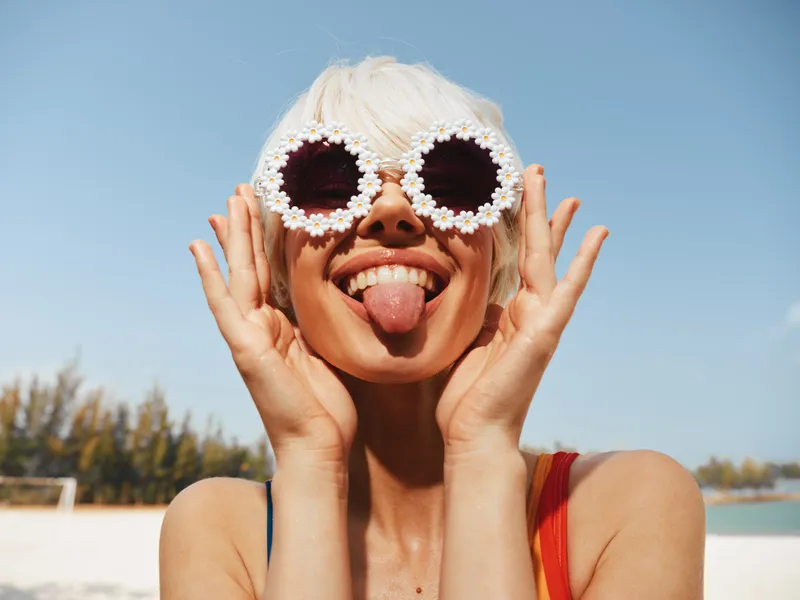 Playful beach portrait of a smiling woman wearing round pearl sunglasses, sticking out her tongue against a bright blue sky and sunlit sand.