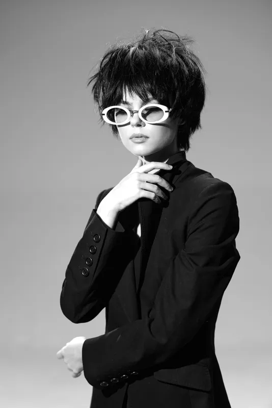 Black and white portrait of stylish woman wearing white sunglasses and dark blazer with thoughtful expression in studio setting.