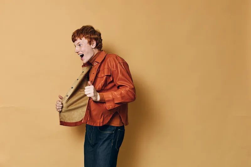 Young man with red hair wearing orange jacket and blue jeans posing excitedly against beige background. Side view, casual clothing and energetic expression in studio setting.