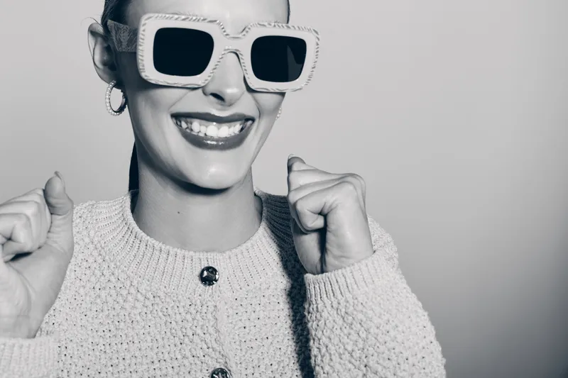 Smiling caucasian woman with light skin wearing oversized sunglasses and knitted sweater poses happily with clenched fists on plain background.