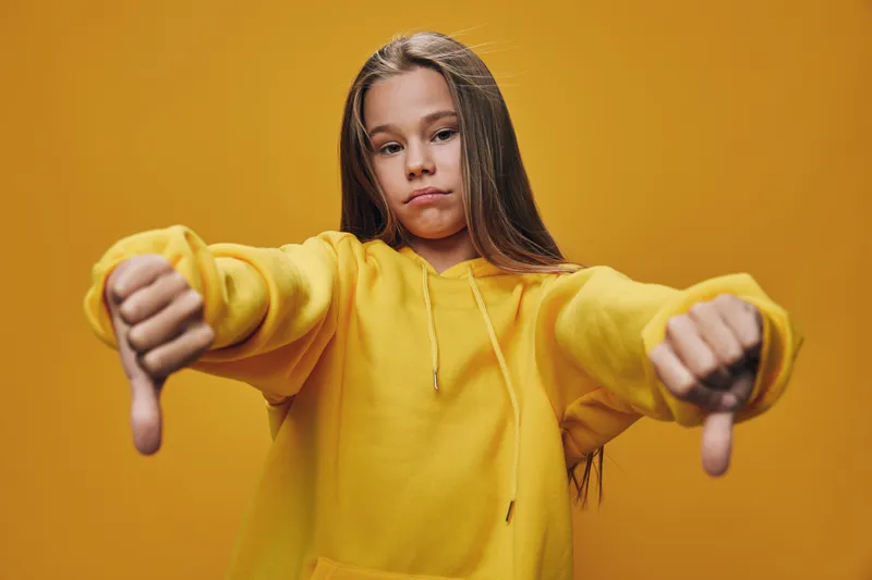Girl in yellow hoodie showing thumbs down with both hands, serious expression on face against yellow background. Child wearing casual clothes, disappointment or disapproval.