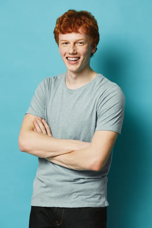 Young man with red hair smiling happily with arms crossed wearing casual gray t shirt and black pants standing against blue background in studio portrait.