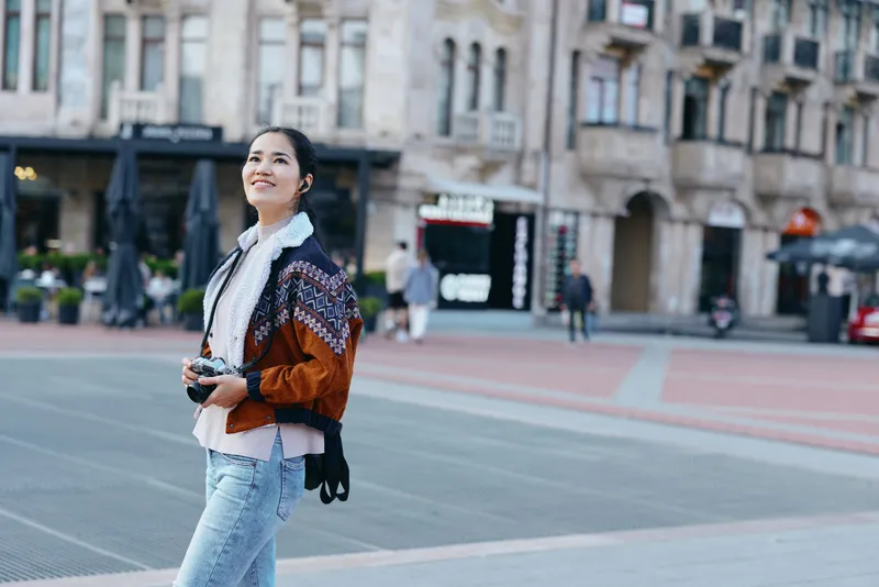 Woman strolling through a city street wearing a knit jacket and scarf, holding a camera outdoors and smiling, urban fashion scene amid historic buildings and pedestrian plaza.