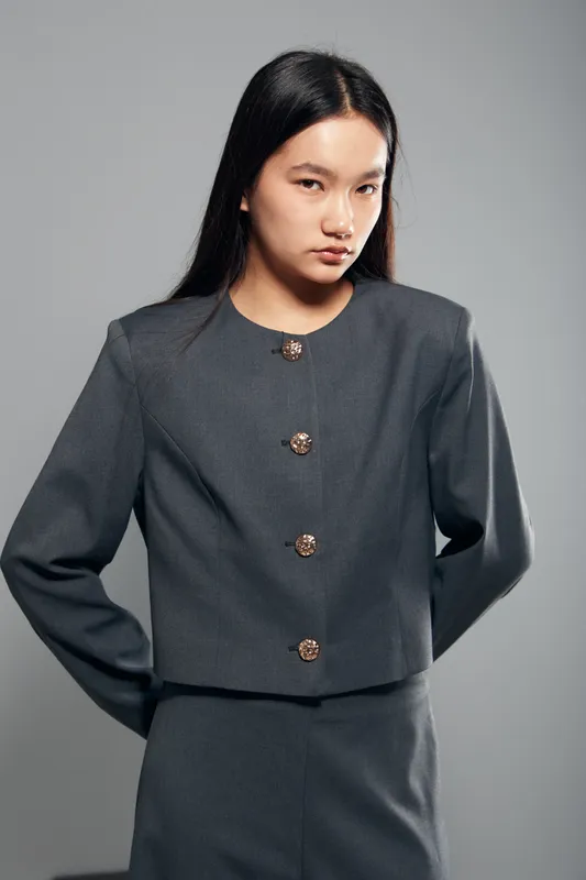 Young woman with long black hair wearing grey formal jacket and skirt posing with serious expression on grey background in studio lighting and minimal makeup fashion style.