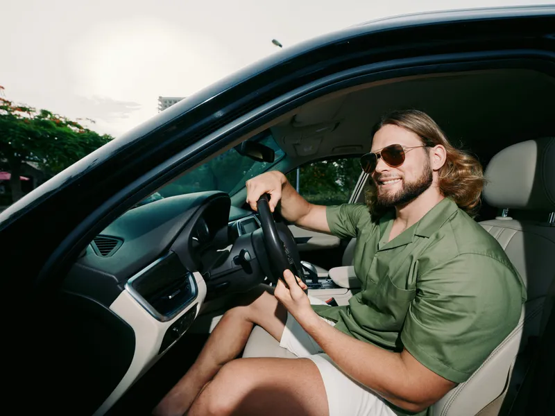 Male driver sits in a modern car, wearing sunglasses and a green shirt, gripping the steering wheel with one hand as sunlight fills the cabin, creating a warm, calm vibe.
