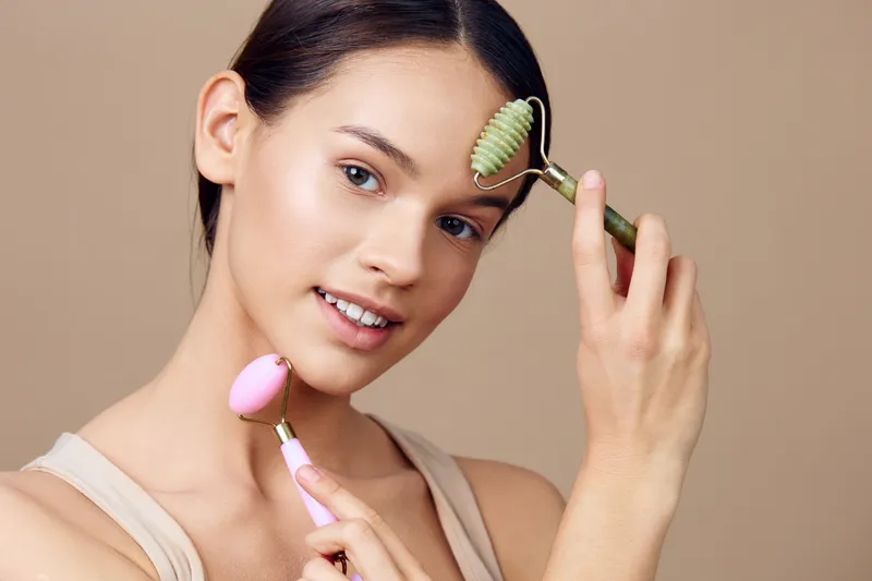 Young woman demonstrating skincare routine with jade roller and gua sha tool on neutral background, promoting healthy skin care concept