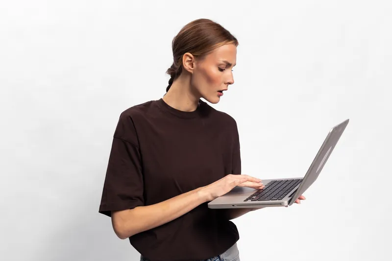Young woman in dark sweater looks surprised while holding laptop with both hands on white background. Concept of technology, communication, and unexpected online news.