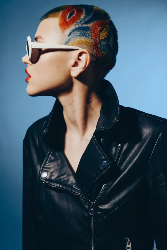 Young woman with colorful shaved hair and geometric tattoo wearing white sunglasses and a black leather jacket against a blue gradient background, showcasing bold style.