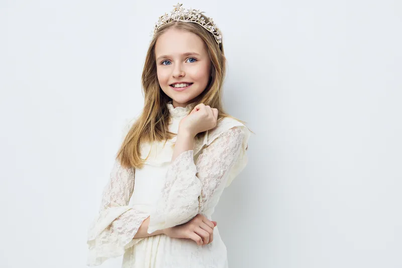 Cheerful girl wearing a crown and elegant dress, posed against a soft white background, showcasing innocence and joy, perfect for children s fashion themes