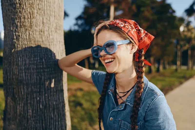 smiling woman with braids wearing sunglasses and red bandana outdoors in warm sunlight enjoying nature and casual blue denim shirt