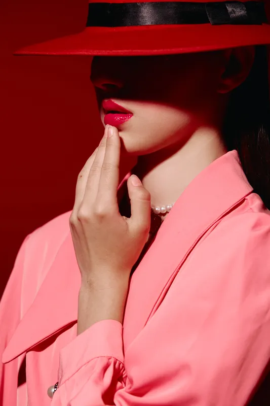 Fashion model embraces creativity with red lips beside shadowed eyes under red hat. Pink trench coat and pearl necklace add cinematic beauty without revealing identity. Bold and mysterious aesthetic.