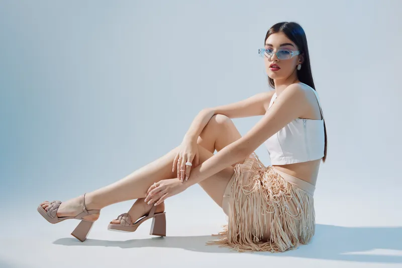 Young woman in stylish beige fringe skirt and white crop top poses seated on white floor. She wears transparent sunglasses and nude heels. Concept of beauty fashion and elegance.