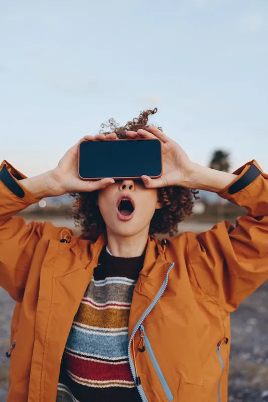 Woman lifestyle outdoors, curly hair, orange jacket, smiling and laughing, holding smartphone in both hands, blue sky background, hands framing the device, casual street moment, natural light