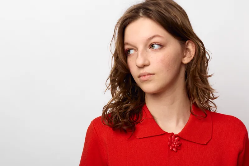 Young woman with wavy brown hair wearing a red sweater looks thoughtfully to the side against a plain white background. Concept of contemplation, casual style, and natural beauty.