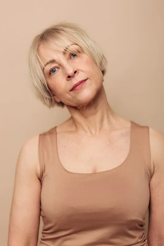 Portrait of a mature woman with short blonde hair in beige top looking thoughtfully at camera against neutral background. Concept of aging beauty, confidence, and natural skin care.