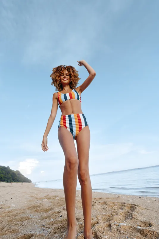 Woman standing on a beach in colorful striped swimsuit, arms up under bright blue sky, ocean waves and sandy shoreline behind, sunny summer vacation mood, relaxed posture, natural colors