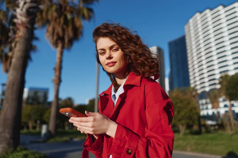 Young woman in a red coat stands outdoors in an urban park, holding a smartphone, with a confident expression and tall buildings in the background.