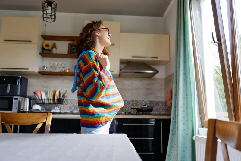 Young woman with curly hair stands in a bright kitchen wearing a colorful striped sweater, looking toward the window with a calm, thoughtful mood