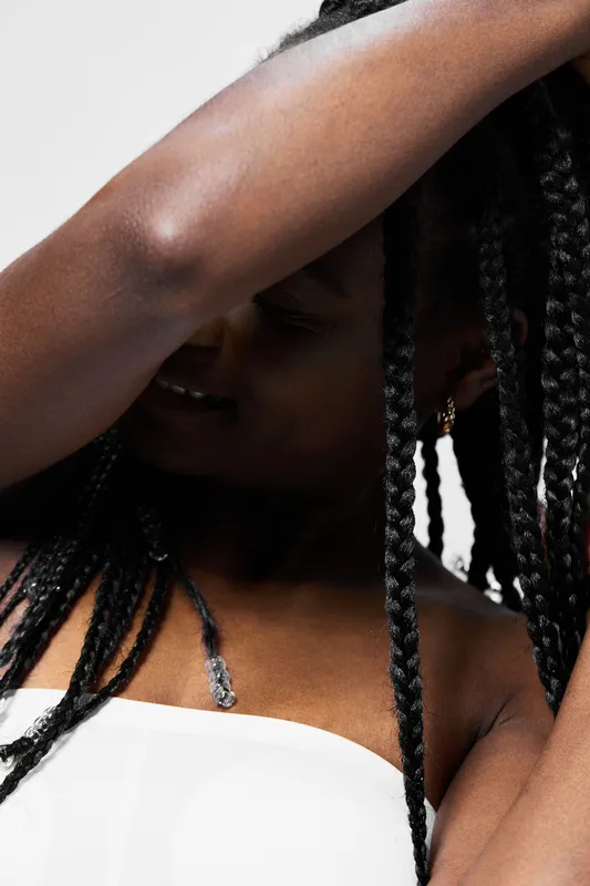 A woman with braided hair playfully adjusts her hairstyle against a bright white background, showcasing self expression and confidence in a modern, contemporary style