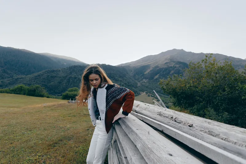Woman leaning on wooden fence in meadow with mountains background, wearing patterned jacket and white pants. Outdoor portrait, countryside landscape and moody natural scenery.