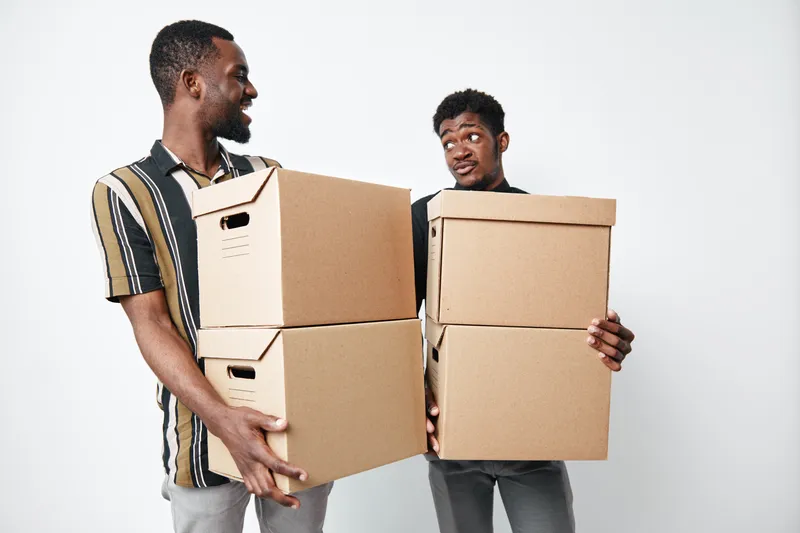 Two african american men carrying cardboard boxes for moving or delivery, smiling and looking at each other on white background. Young black men with dark skin holding packages.