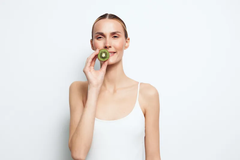 Young woman with a brunette bun wears a white tank top and bites a kiwi against a clean white background, presenting a fresh, healthy snack moment.