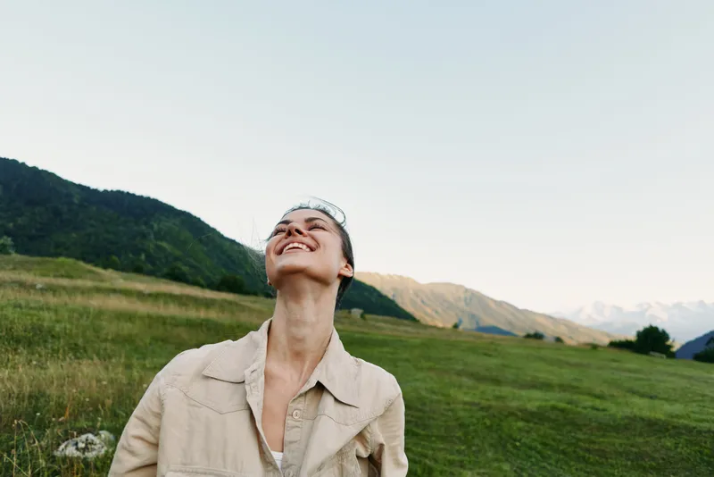A young woman in a beige jacket tilts her head back and smiles in a sunlit meadow under a clear sky, capturing a moment of lighthearted outdoor freedom and contentment.