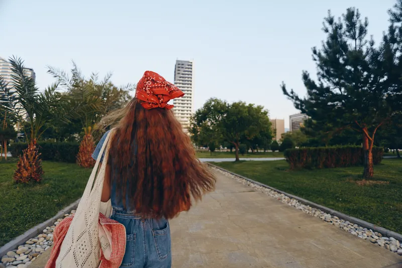 Lifestyle woman in modern boho-western denim street style with long hair and red bandana walking in park under clear sky, film color tones enhance casual vibe.