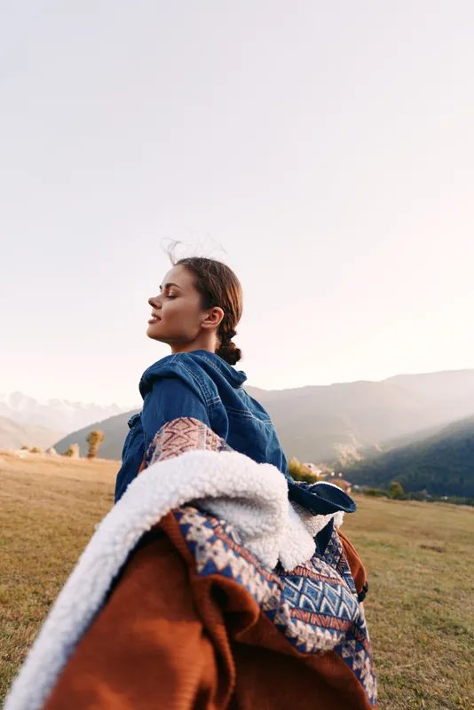 Woman holding hand in a grassy field with blanket and denim jacket, smiling against distant mountains. Travel adventure and outdoor couple moment capturing carefree exploration.