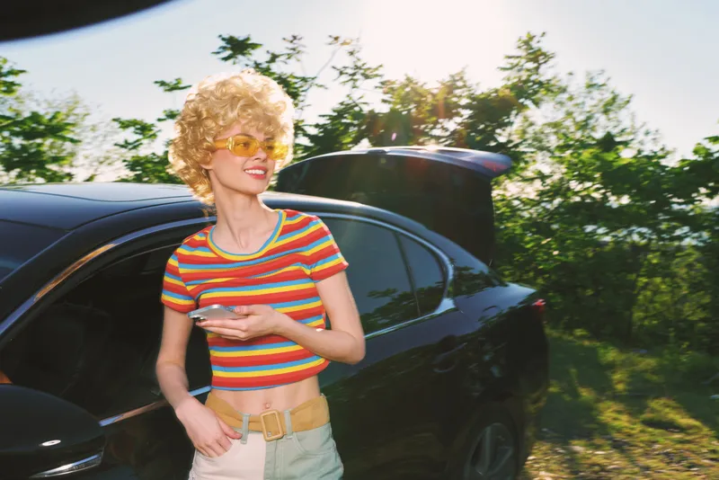 Smiling woman with curly hair beside a sleek car in bright sunlight. Casual fashion, outdoor lifestyle shot with retro film color mood and carefree summer vibe.