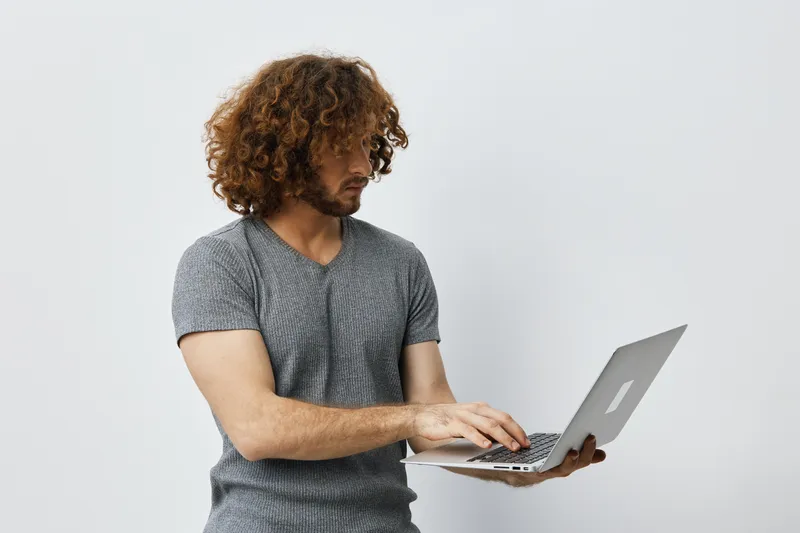 Young man with curly hair working on a laptop, showcasing concentration in a minimalistic, light background perfect for technology and education concepts