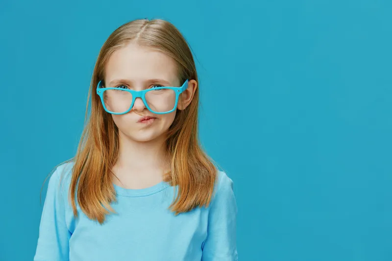 Young girl with blue glasses making a funny face against a solid blue background, showcasing playful emotions in a vibrant and colorful setting