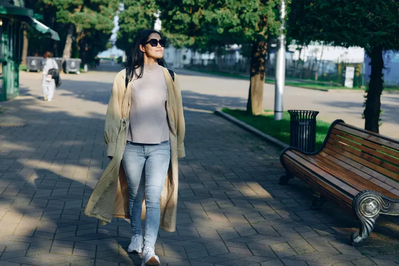 Woman strolling along a sunny park street in a long coat, casual chic style, sunglasses and relaxed confidence, showcasing everyday fashion and outdoor lifestyle in motion and calm surroundings