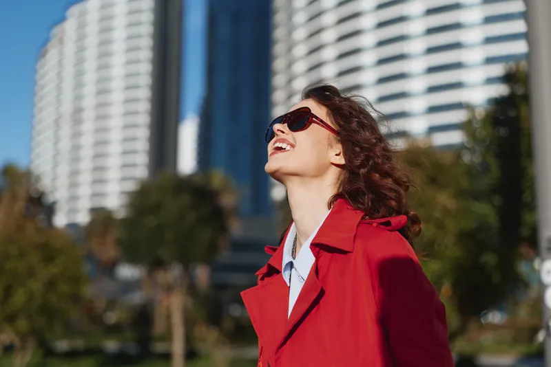 Young woman in a red coat and sunglasses smiles joyfully while looking up outdoors in an urban park with modern buildings in the background on a sunny day.