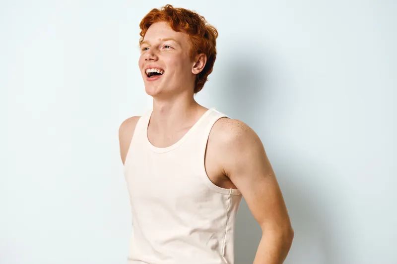 Young man with red hair wearing white tank top smiling and laughing on white background. Happy male portrait with casual sleeveless shirt and positive expression.